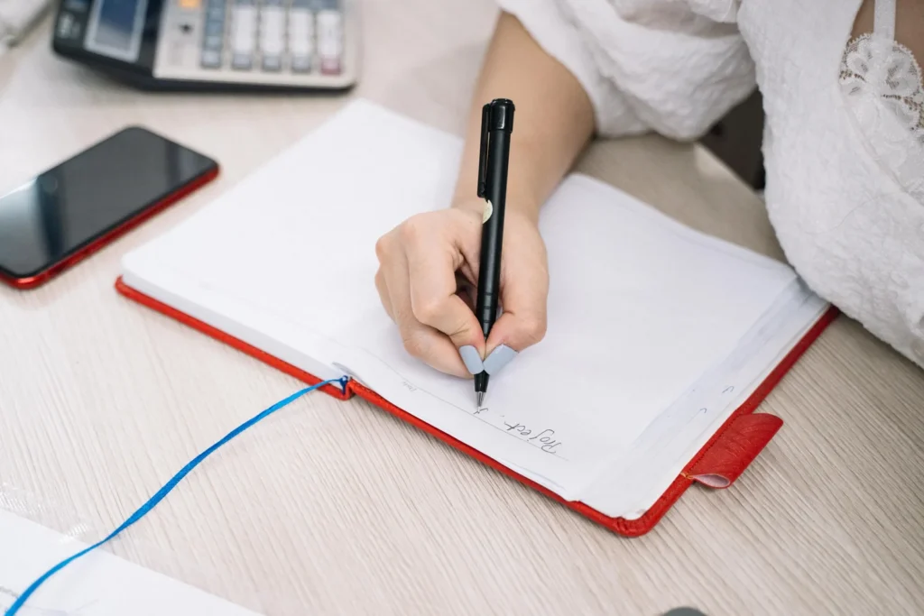 A hand writing notes in a notebook on a desk beside a phone