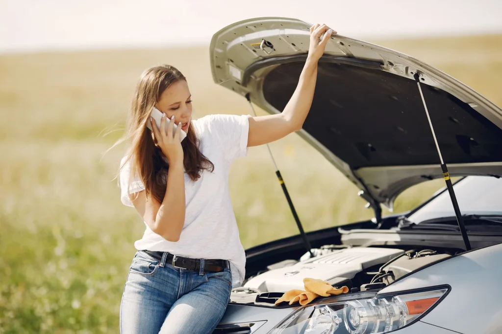 A worried driver leaning on her car with the hood open, calling for help from the roadside
